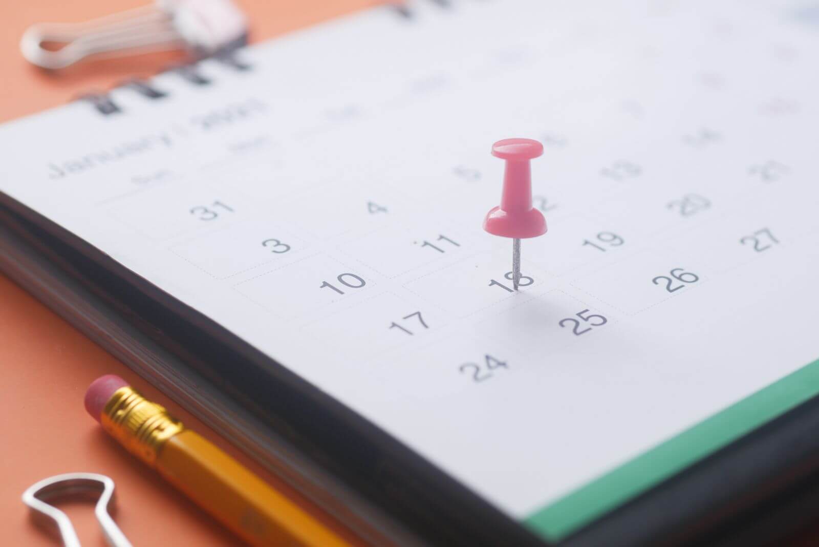 Coaching: A close-up of a calendar page with a pink push pin, pencil, and binder clip on an orange background.